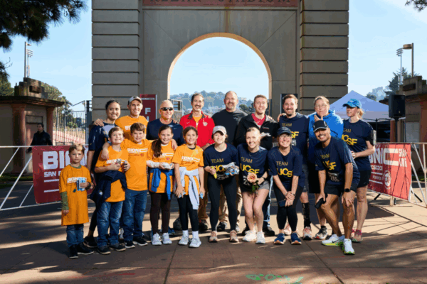 A large group of adults and children wearing event shirts pose together for a group photo in front of the Kezar Stadium archway after the event.