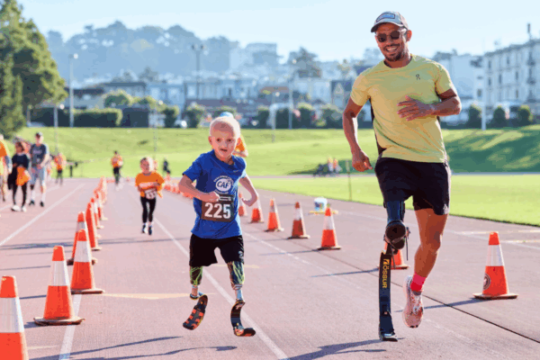 A young boy with running blades runs down the track beside an adult athlete using a running blade, with event cones and other participants in the background.