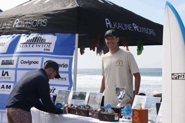 A Surf for Impact volunteer stands smiling beside the event prize table, displaying trophies and giveaway items under an Alkaline Advisors tent with the ocean in the background.