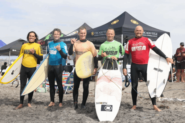 Five surfers in colored competition jerseys stand on the beach holding their surfboards, smiling and giving shaka signs in front of event tents.