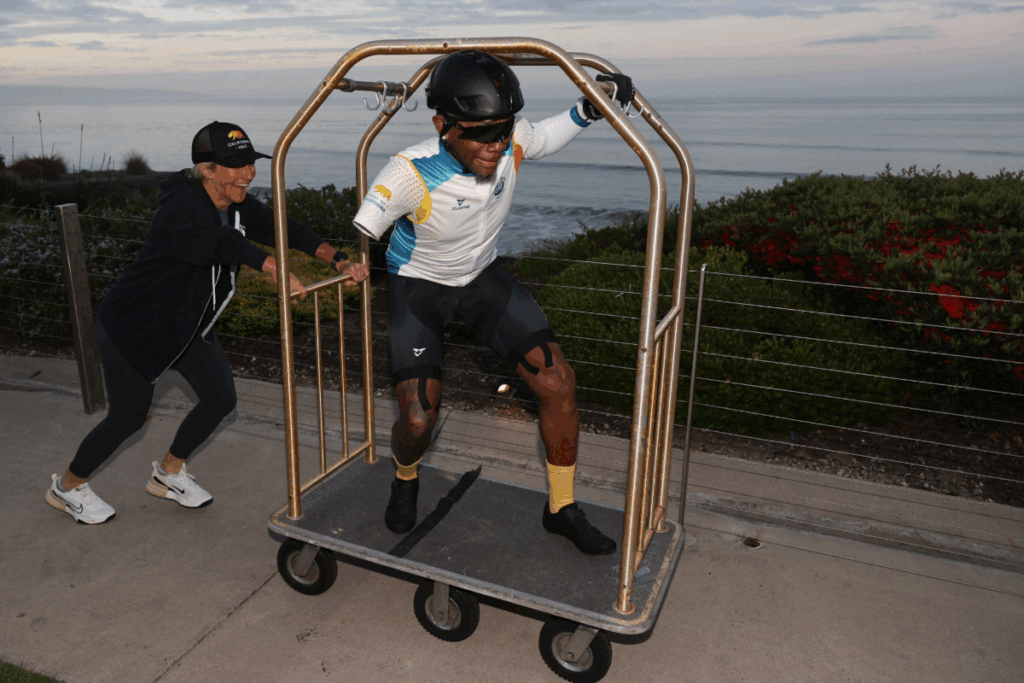 Athlete with a prosthetic leg laughing while being pushed on a hotel luggage cart near the ocean.