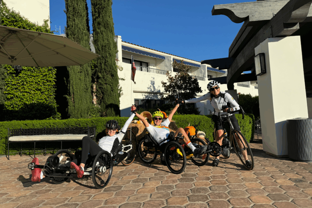 Three athletes using handcycles and a road bike smiling outside a hotel courtyard decorated with pumpkins.