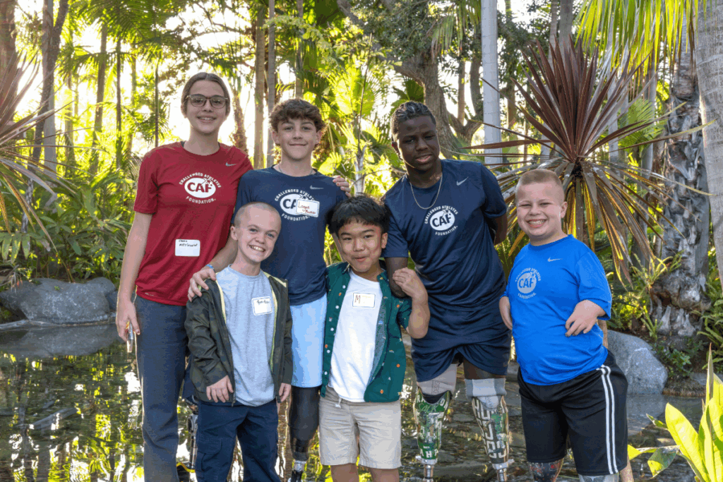 Group of six young athletes, some with prosthetic legs, smiling together in a tropical garden setting.