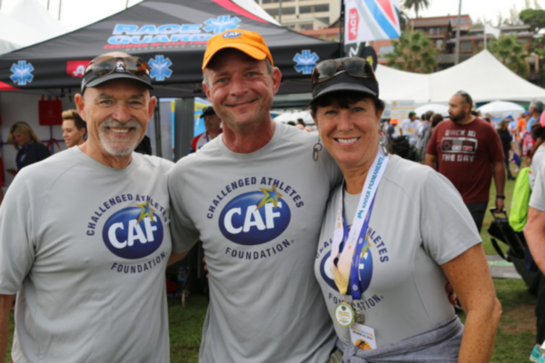 Three adults wearing Challenged Athletes Foundation shirts stand arm in arm at a race village, smiling after completing an event, with tents and other participants in the background.