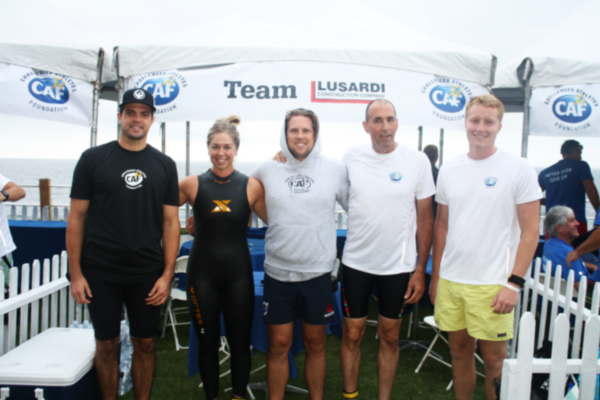 Five athletes stand shoulder to shoulder in front of a Team Lusardi and CAF tent near the ocean, smiling after a race, with wetsuits and athletic gear visible.