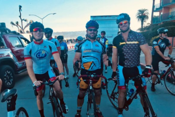 Three cyclists wearing helmets and CAF cycling jerseys pause with their bikes at the start of a ride, smiling as other riders gather behind them along a coastal road.