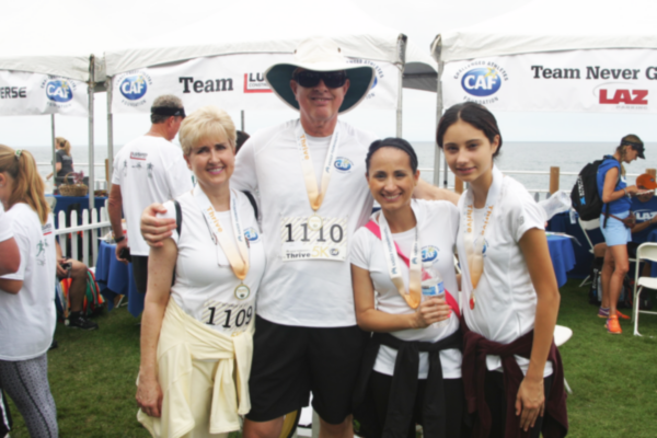 Four participants wearing CAF shirts and finisher medals stand together near the ocean, smiling and embracing after completing a 5K event.