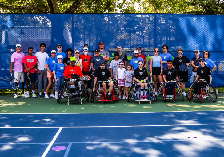 Group photo of athletes, volunteers, and coaches gathered on an outdoor tennis court during an adaptive tennis clinic.