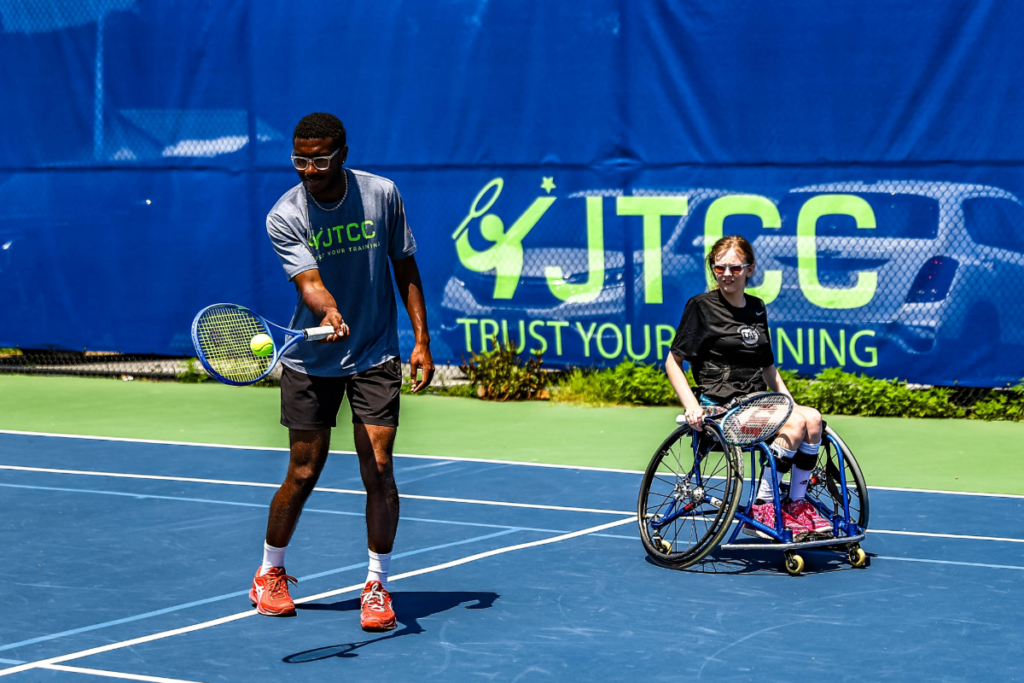 Two athletes participate in an adaptive tennis drill on an outdoor court, with a coach demonstrating ball control beside a wheelchair tennis athlete.