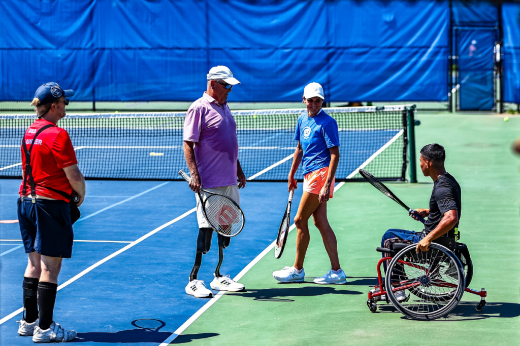 Athletes and a coach pause between points on an outdoor tennis court during an adaptive tennis clinic, holding rackets and smiling.