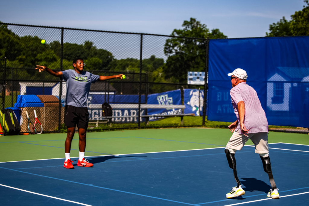 Athletes practice adaptive tennis on an outdoor court as a coach tosses balls during a skills exercise.