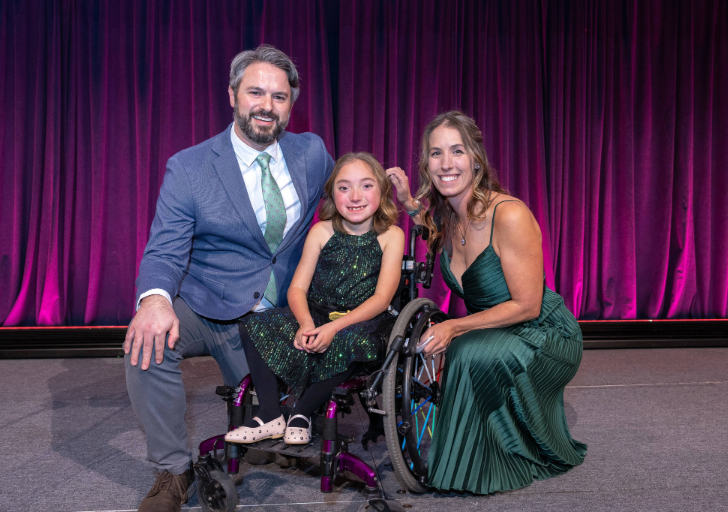A young girl using a wheelchair smiles on stage between two adults in formal attire, with a purple curtain backdrop behind them at the Heroes, Heart & Hope Gala.