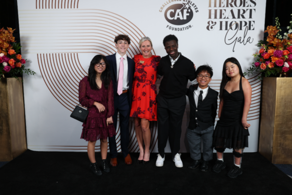 A group of six young people in formal attire pose together in front of the Heroes, Heart & Hope Gala step-and-repeat, smiling in front of floral arrangements and CAF branding.