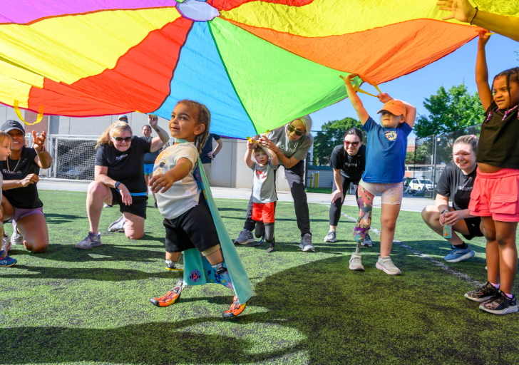 Children with physical disabilities and volunteers play together under a colorful parachute during an inclusive outdoor activity.