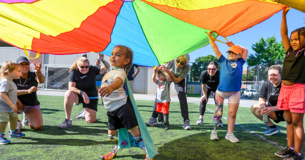 Children with physical disabilities and volunteers play together under a colorful parachute during an inclusive outdoor activity.