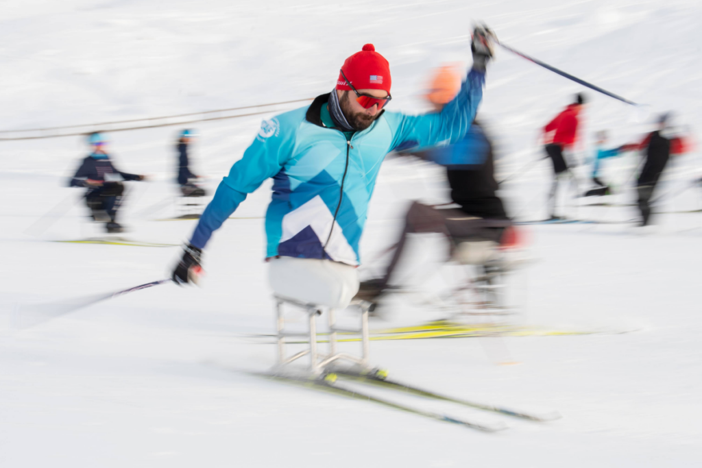 A person wearing a blue jacket and red hat uses a sit-ski on a snowy course, captured in motion with other sit-skiers and participants blurred in the background.