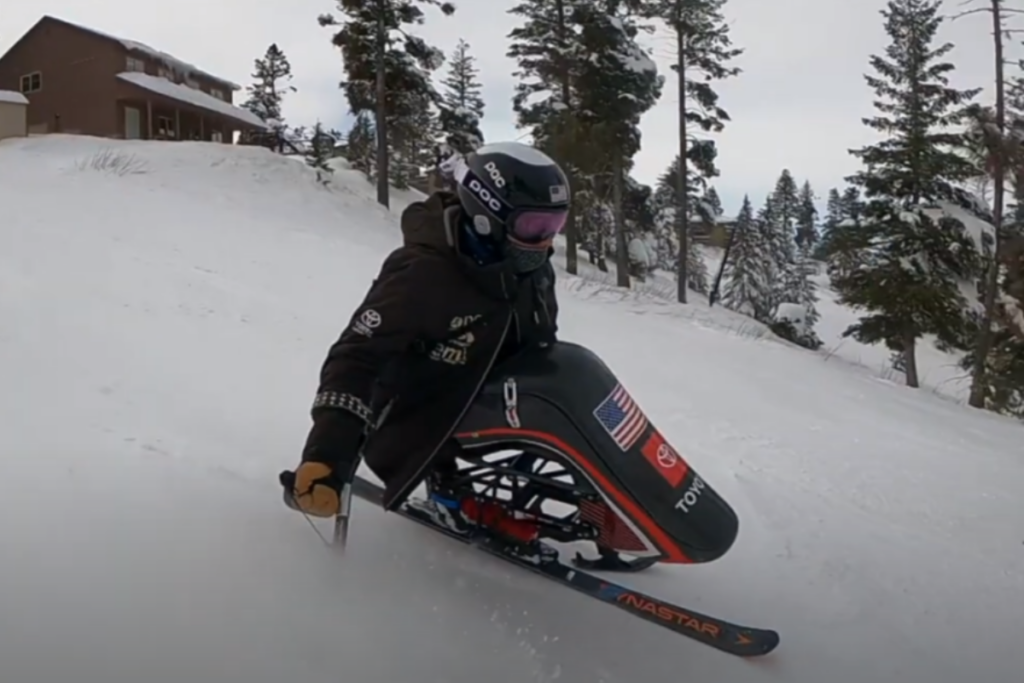 A person wearing a helmet and goggles navigates a sit-ski down a snowy slope, with pine trees and a building in the background.