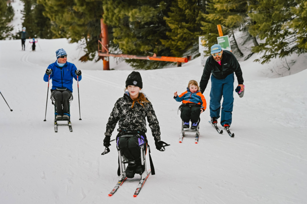 A group of people using sit-skis moves together on a snowy trail, accompanied by an instructor or guide. Evergreen trees and snow-covered ground are visible in the background.