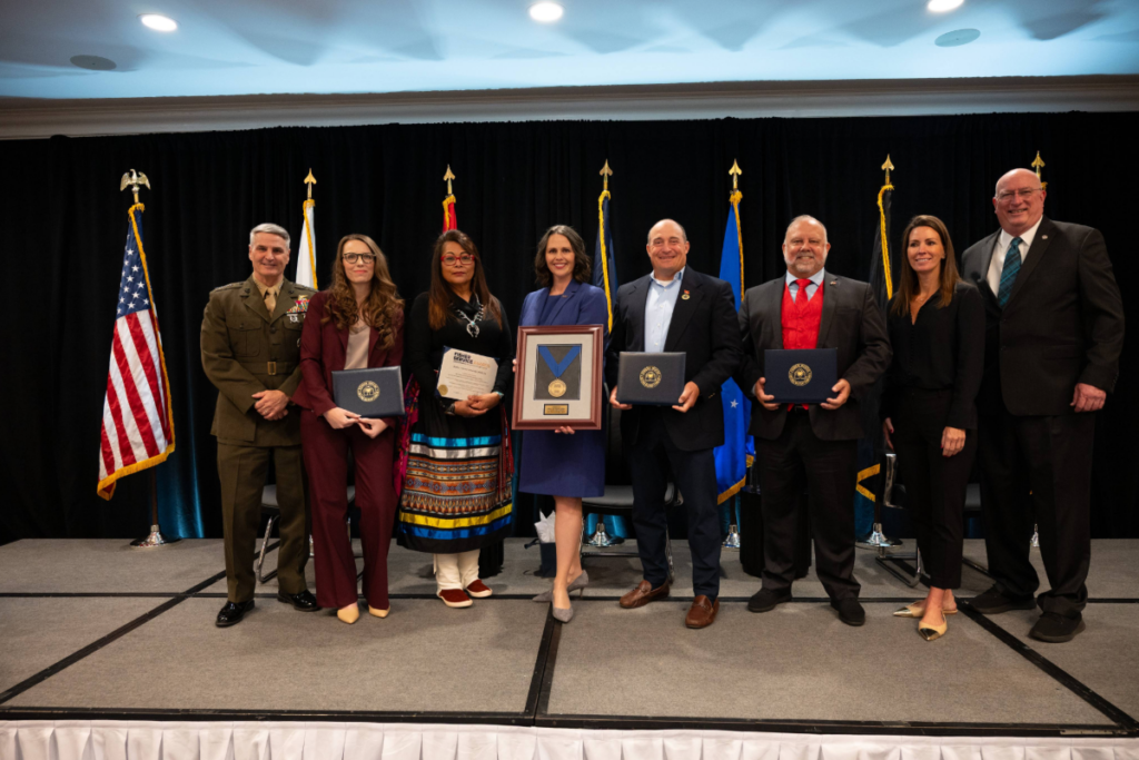 Fisher Service Award recipients stand on stage holding certificates and medals, framed by U.S. military service flags.