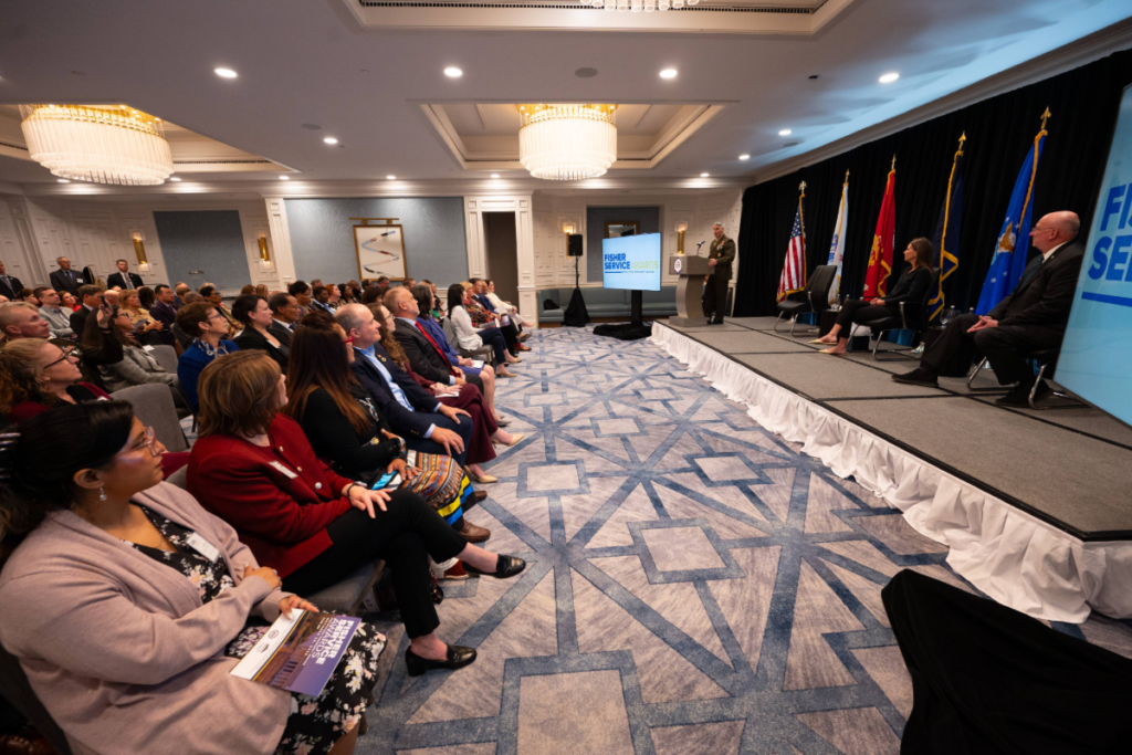 Audience listens as a speaker presents at the Fisher Service Awards ceremony, with service branch flags and large screens on stage.
