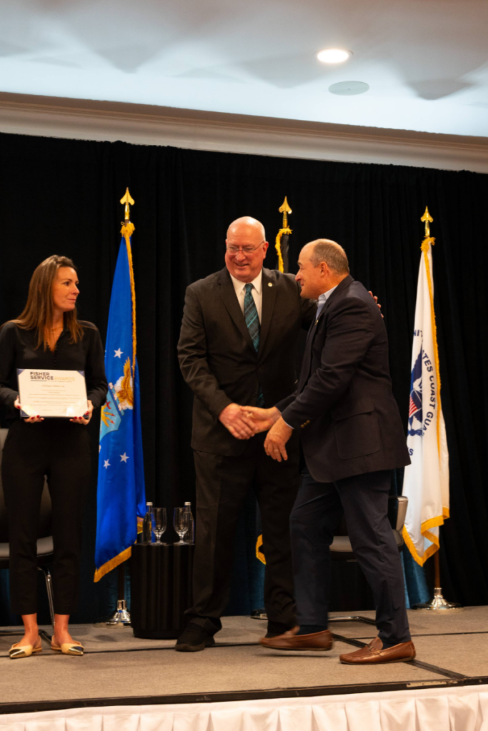 A Fisher Service Award recipient shakes hands with a presenter on stage as another presenter holds the award certificate.