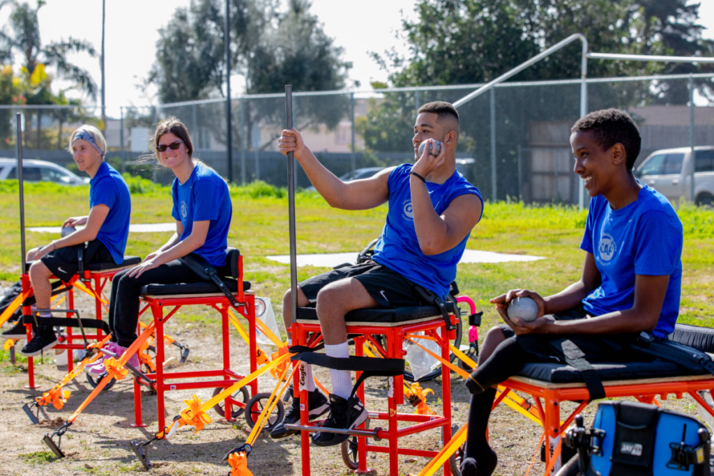 Teen athletes with physical disabilities participate in a seated throwing activity outdoors, holding shot put implements and training equipment.