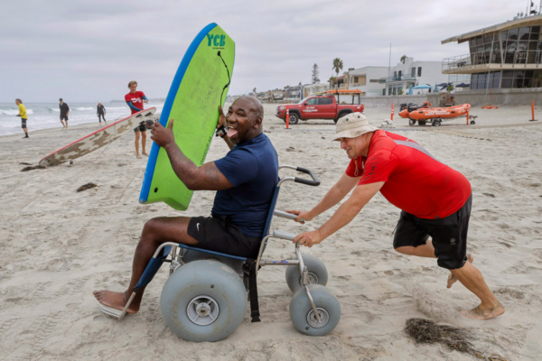 An athlete with a physical disability sits in a beach wheelchair holding a surfboard while a volunteer pushes them across the sand toward the water at a CAF Operation Rebound surf clinic.
