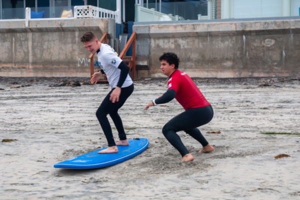 An athlete practices standing on a surfboard on the sand while a volunteer supports from behind during a CAF Operation Rebound surf clinic.