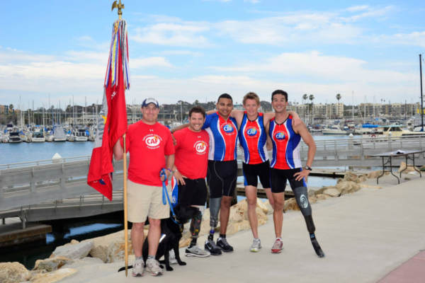 A group of CAF athletes with physical disabilities and supporters stand together by a marina, wearing CAF triathlon gear and smiling after a community event.