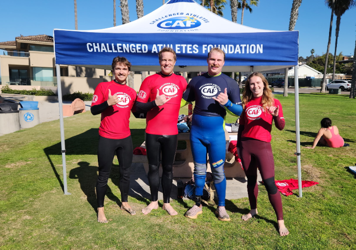 Athletes with physical disabilities stand barefoot on the grass after a surf session, wearing CAF rash guards and wetsuits, smiling and giving shaka signs beneath a Challenged Athletes Foundation tent at La Jolla Shores.