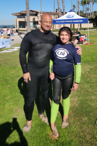 An adult mentor and a young athlete with a physical disability stand side by side on the grass after surfing, wearing CAF wetsuits and rash guards, smiling in front of a Challenged Athletes Foundation tent near the beach.