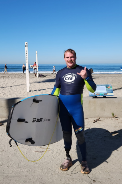 An athlete with a physical disability stands on the sand holding a surfboard after a session, wearing a CAF rash guard and wetsuit, smiling and giving a shaka with the ocean visible behind them.