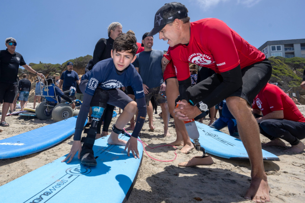 A coach kneels beside a young surfer with a lower-limb prosthesis, offering guidance as the athlete prepares to enter the water.