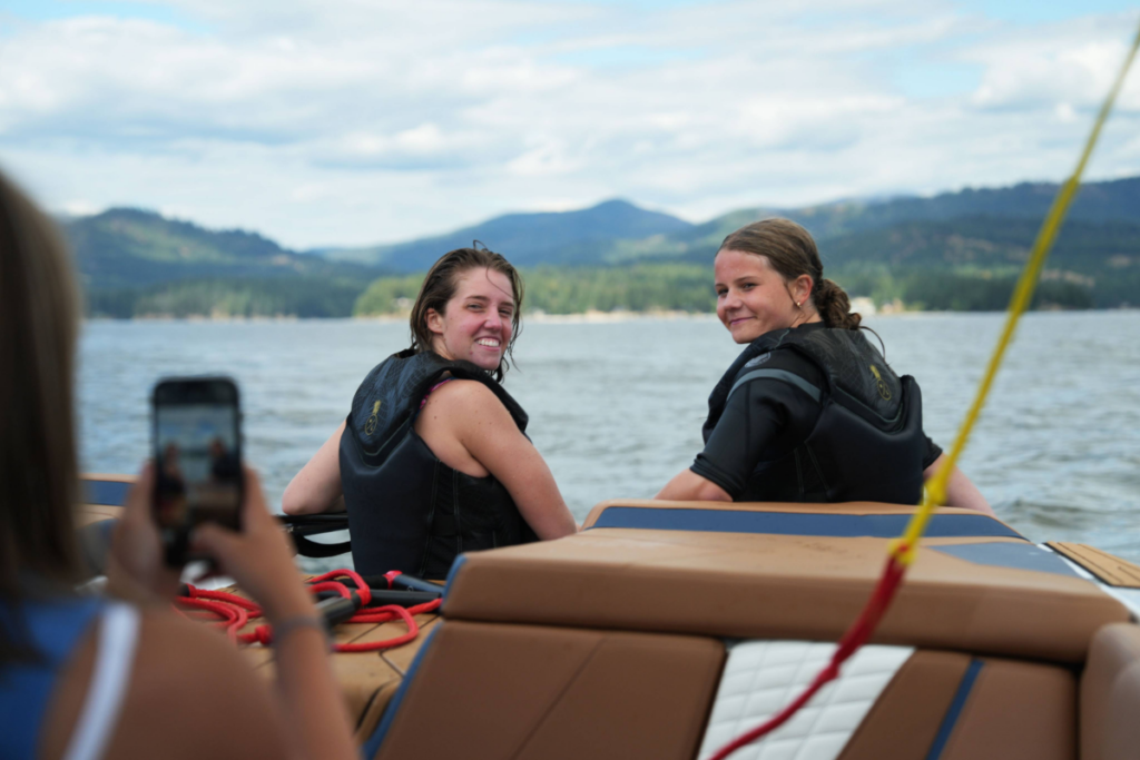 Two people seated on a boat wearing life vests look back toward the camera with a lake and mountains in the background.