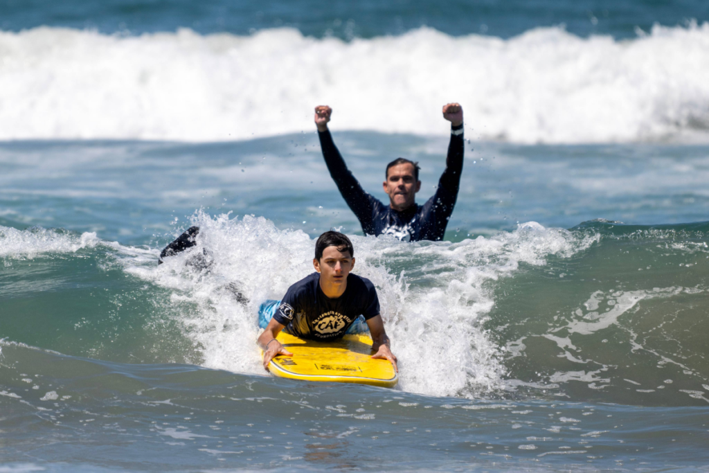 A young athlete rides a wave on a bodyboard while a coach raises their arms in celebration behind them.