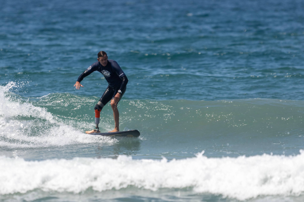 A surfer with a lower-limb prosthesis rides a small wave while wearing a CAF rash guard.