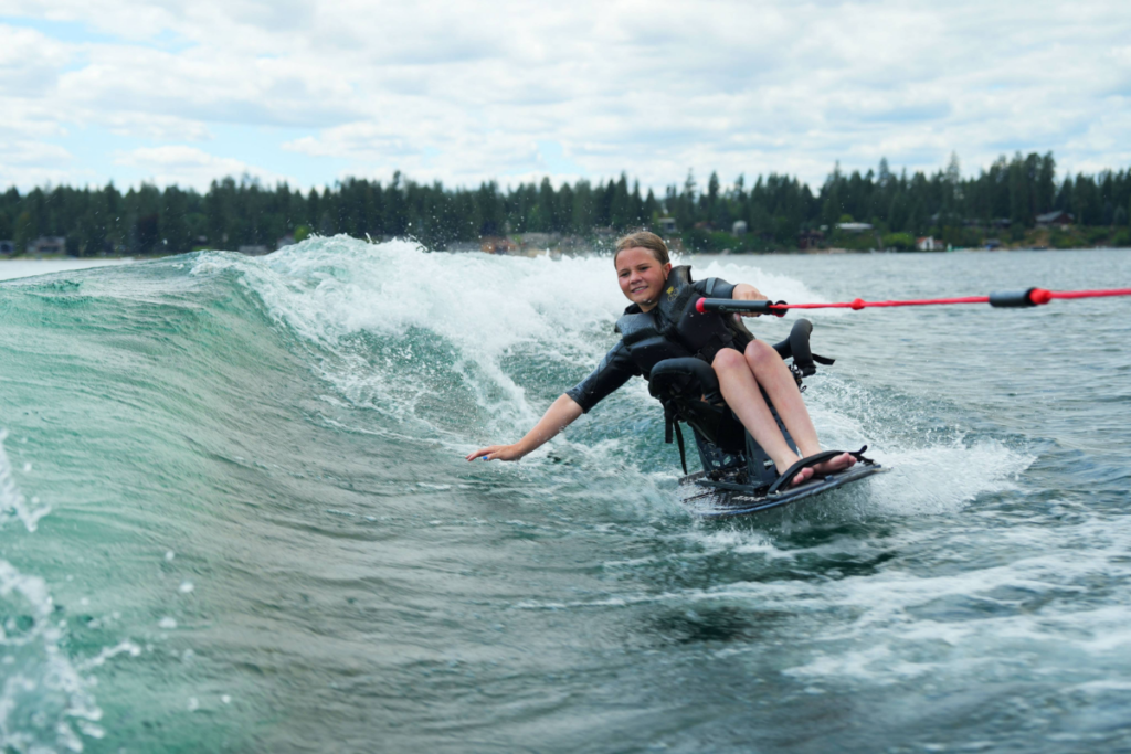 A young person wake surfing in an adaptive seat skims the surface of a lake, reaching one hand toward the water.