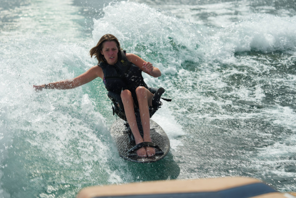 A wake surfer in an adaptive seat carves through a wave with water splashing around the board.