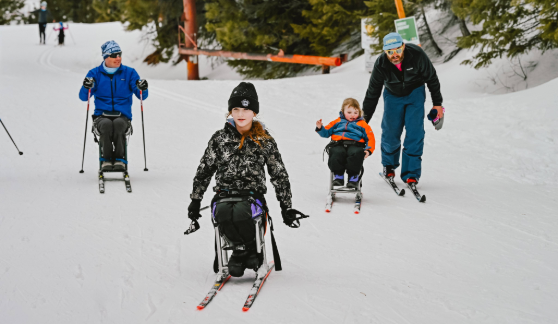 A group of athletes using sit-skis gliding down a snowy trail, accompanied by a coach or volunteer. Evergreen trees and snow-covered ground are in the background.