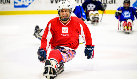 A sled hockey player in a red jersey moving across the ice, with other players in blue jerseys behind. The scene takes place in an indoor ice rink.