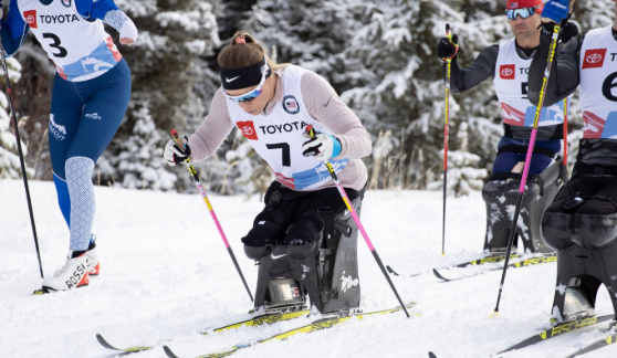 Sit-ski athletes racing on a snowy course, each using poles to propel forward. The event is set outdoors with snow-covered trees in the background.
