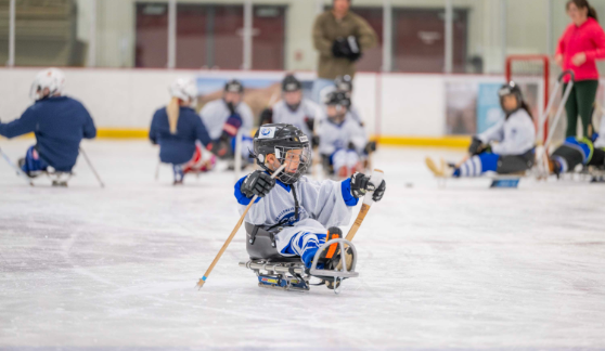 A young athlete playing sled hockey on an ice rink, using sticks to propel forward. Other players and coaches are visible in the background.