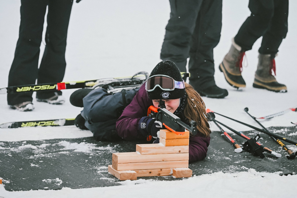 A para biathlete lying on the snow, aiming a rifle at a target during a winter sports event. Cross-country skis and poles are visible nearby, with other participants standing in the background.