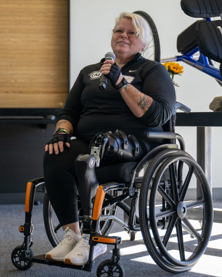 A person in a wheelchair with prosthetic legs speaks into a microphone, with a bicycle in the background and flowers attached to the chair.