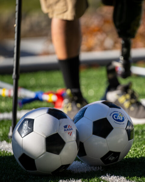 Two soccer balls on a green turf field with a person's legs, one prosthetic, behind them.