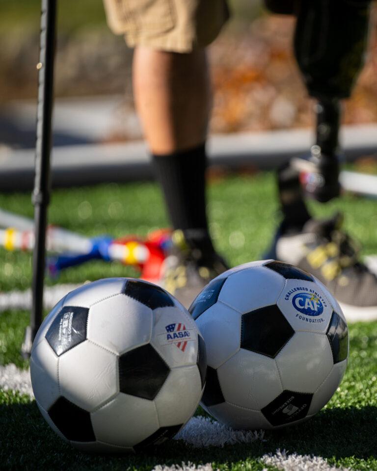 Two soccer balls on a green turf field with a person's legs, one prosthetic, behind them.