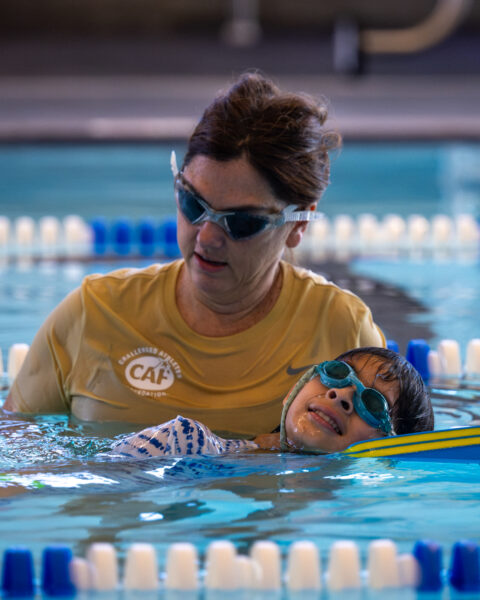 A person helps a child in a blue vest and goggles in a swimming pool.