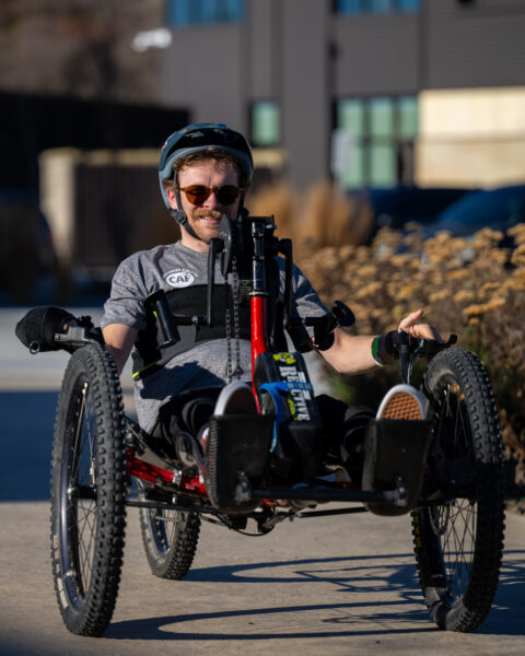 Person riding a recumbent handcycle on a sidewalk with a building in the background.