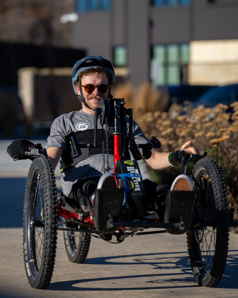 Person riding a recumbent handcycle on a sidewalk with a building in the background.