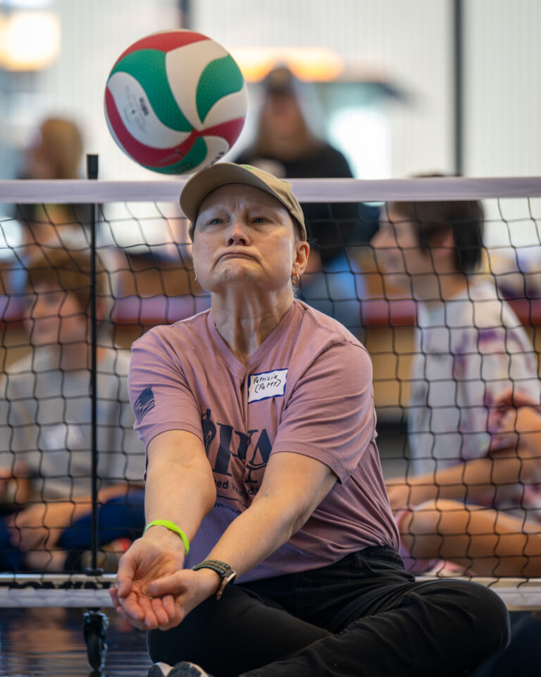 Person playing sitting volleyball, focused on receiving a colorful volleyball above a net.